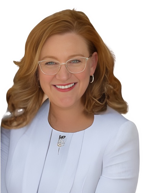 Headshot of Alysia Cook, a smiling woman with shoulder-length wavy reddish-brown hair, wearing glasses and a white blazer against a simple background.