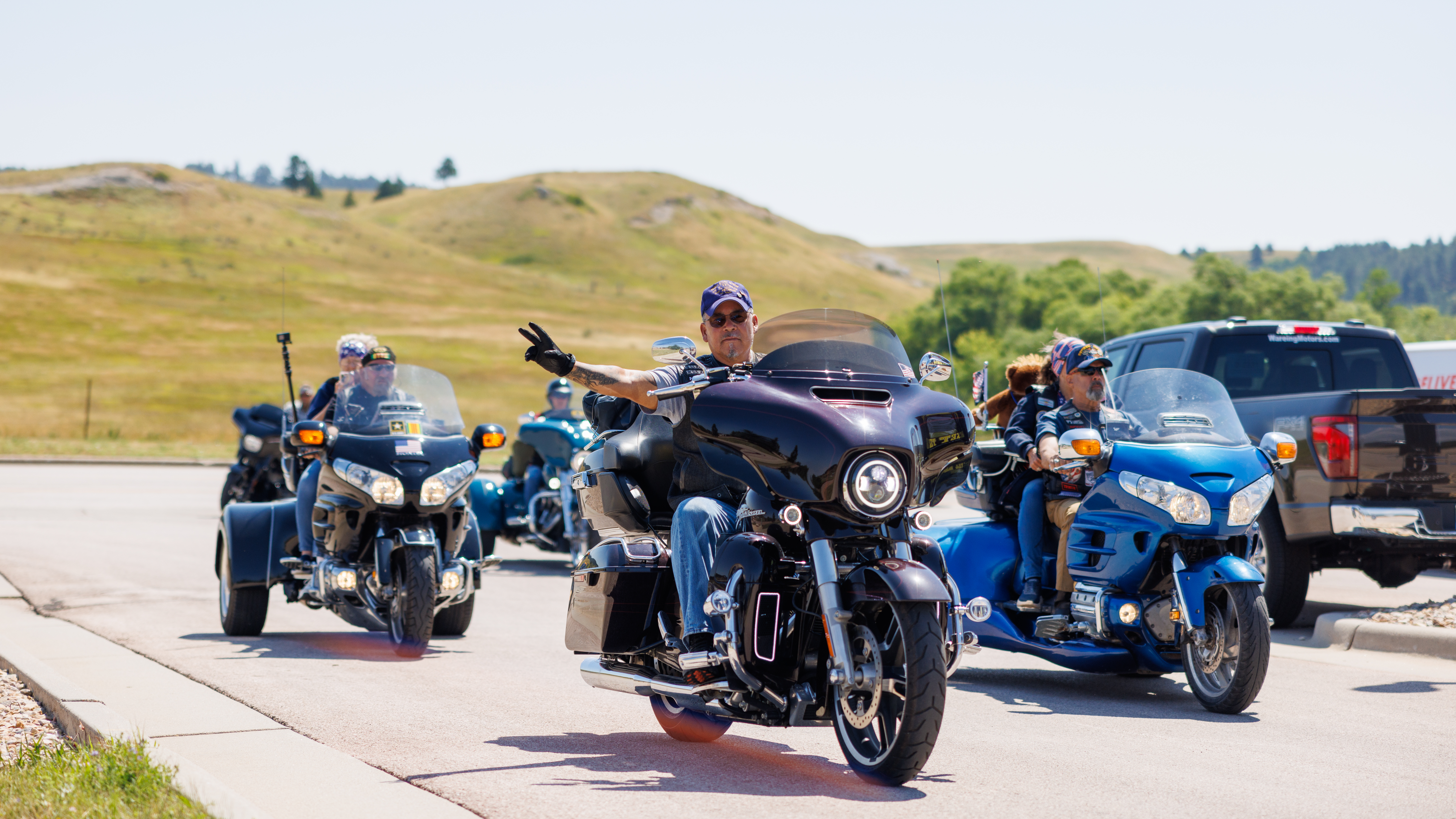 A group of motorcyclists on their bikes in a parking lot with rolling hills in the background; the lead rider gives a peace sign to the camera.
