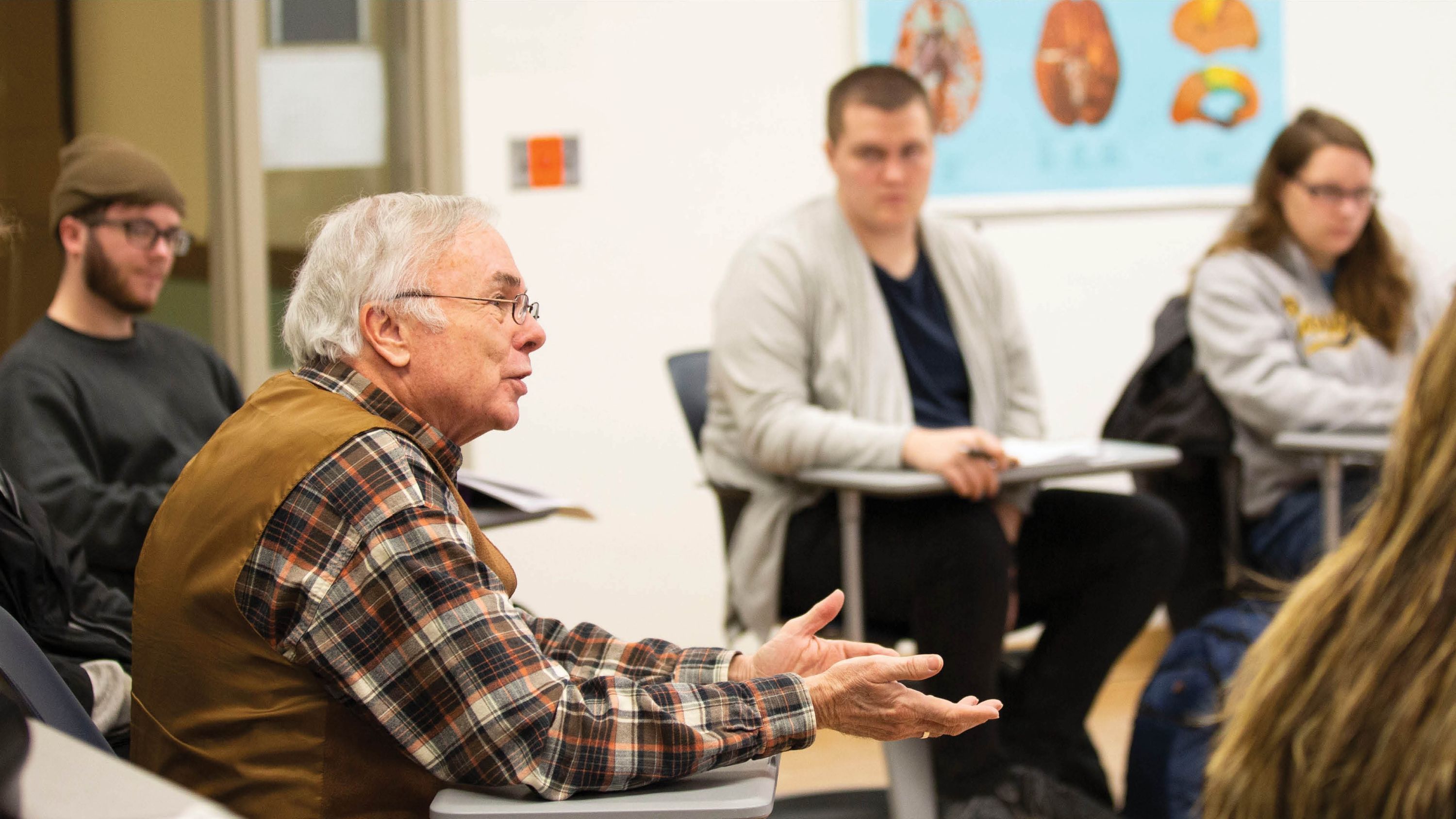 A male professor speaks during a classroom discussion while students listen and take notes.