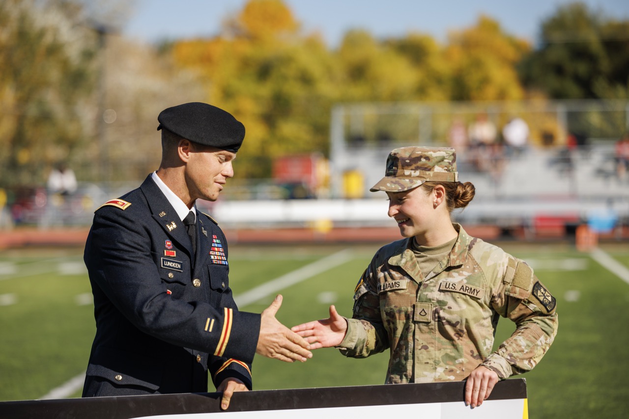 A military-affiliated graduate smiles while holding his diploma, wearing a black gown with gold honors sash. A crowded gym with fellow graduates in yellow robes shows how BHSU supports both veterans and active military in achieving their education goals.
