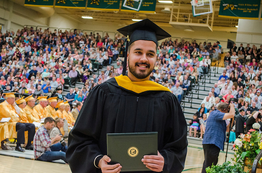 Picture of BHSU graduate holding his diploma; many other BHSU graduates sitting in the background with an audience sitting in the bleachers in the Young Center Gym.